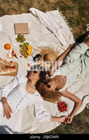 vista dall'alto di ragazze afroamericane positive che si tengono per mano e si posa su una coperta durante il picnic Foto Stock