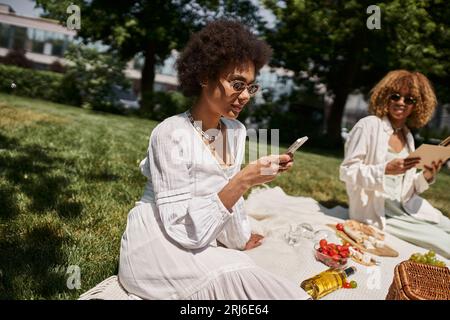 donna afroamericana che utilizza lo smartphone durante il picnic estivo con la ragazza Foto Stock