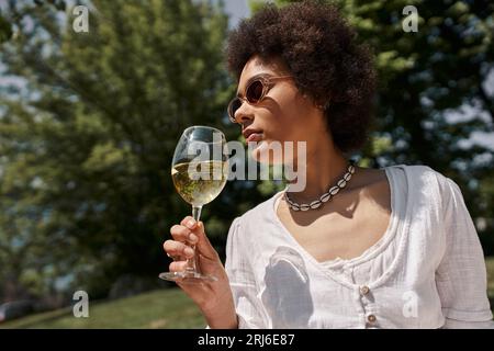 donna afro-americana alla moda con occhiali da sole che beve vino durante un picnic nel parco Foto Stock