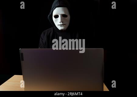 hacker maschile vestito di nero con un cappuccio e una maschera bianca dietro un computer Foto Stock