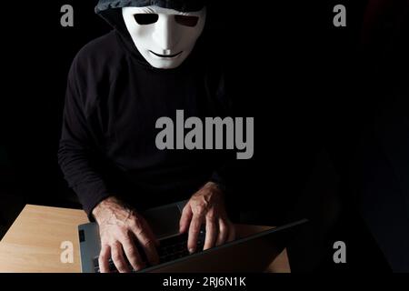 hacker vestito di nero, con cappuccio e maschera che digita su un concetto di cybercrimine Foto Stock