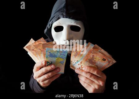 hacker vestito di nero con maschera bianca e cappuccio nero che conta le banconote in euro nelle sue mani, concetto di cybercrimine, isolato su sfondo nero Foto Stock