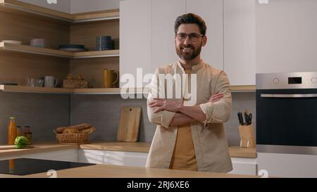 Foto in movimento uomo caucasico adulto con gli occhiali a casa cucina moderna che posa le mani incrociate sorridendo alla macchina fotografica. Ritratto 40 uomo padrone di casa Foto Stock