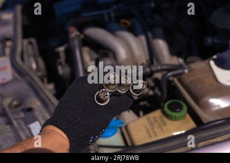 Primo piano di una mano in un guanto nero con quattro candele usate per auto sullo sfondo del motore. Concetto di riparazione auto Foto Stock