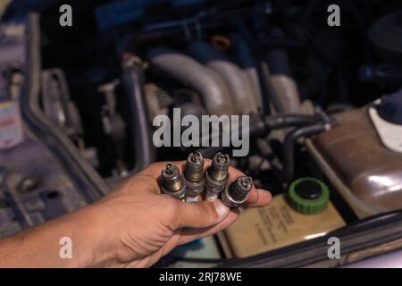 Primo piano di una mano con quattro candele usate sullo sfondo del motore. Concetto di riparazione auto Foto Stock