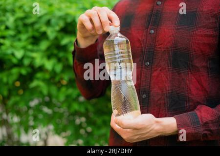 Un uomo irriconoscibile con una camicia rossa tiene tra le mani una bottiglia d'acqua trasparente in primo piano su uno sfondo verde naturale Foto Stock