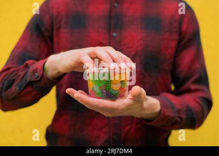 Primo piano uomo irriconoscibile in camicia rossa con caramelle colorate in una scatola di plastica trasparente con sfondo giallo Foto Stock