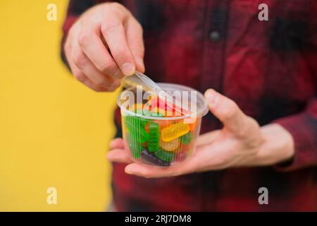 Primo piano uomo irriconoscibile in camicia rossa con caramelle colorate in una scatola di plastica trasparente con sfondo giallo Foto Stock