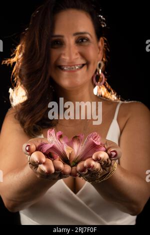 Ritratto in studio di una bella donna matura, che indossa un vestito bianco, con fiori in mano. Isolato su sfondo nero. Foto Stock