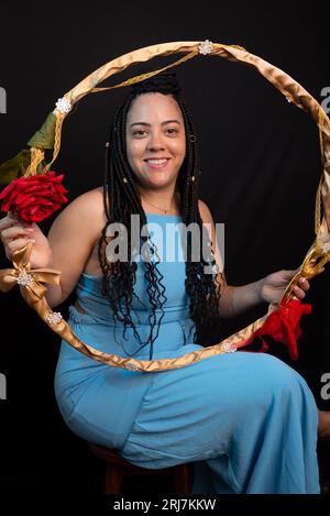 Bella donna che indossa un abito di colore blu seduta con un hula hoop su sfondo nero in studio. Persona positiva. Foto Stock