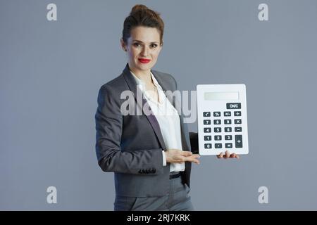 lavoratrice di mezza età felice e alla moda in tuta grigia con calcolatrice isolata su sfondo grigio. Foto Stock