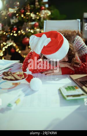 Natale. Donna d'affari stanca ed elegante con cappello da babbo Natale e maglione rosso natalizio con documenti che dormono in un moderno ufficio verde con albero di Natale Foto Stock