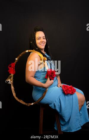 Bella donna che indossa un abito di colore blu seduta con un hula hoop su sfondo nero in studio. Persona positiva. Foto Stock