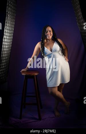 Ritratto in studio di una bella donna con capelli intrecciati in abito bianco che posa per una foto, in piedi. Isolato su sfondo blu scuro. Persona positiva Foto Stock