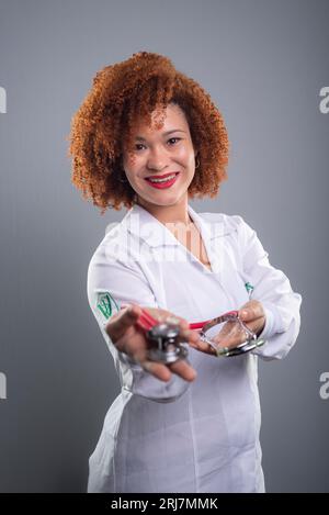 Donna veterinaria, bella con i capelli rossi, che mostra uno stetoscopio alla macchina fotografica in uniforme bianca. Isolato su sfondo grigio. Foto Stock