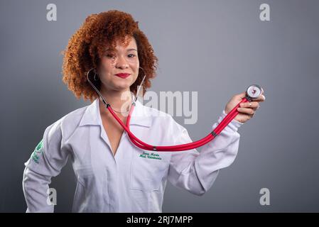 Donna veterinaria, bella con i capelli rossi, che mostra uno stetoscopio alla macchina fotografica in uniforme bianca. Isolato su sfondo grigio. Foto Stock