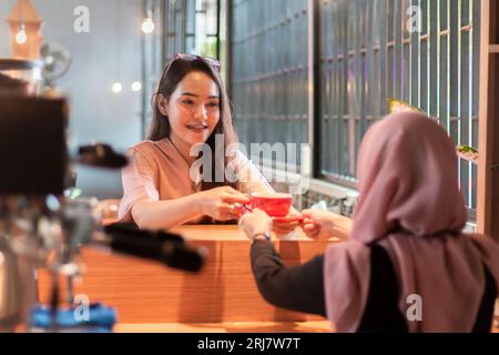 Un bellissimo barista asiatico che consegna una tazza di caffè ai clienti che aspettano gli ordini al bar Foto Stock