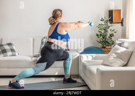 Donna in sovrappeso che fa esercizio sportivo in soggiorno. Affonda e tiene i manubri in mano. Foto Stock