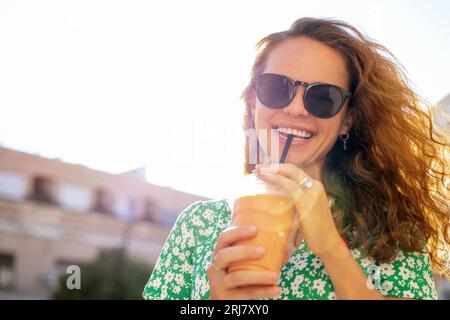 Giovane donna urbana con i capelli castani ricci sta in piedi su strada bevendo succo di frutta usando cannuccia e sorridendo. Foto Stock