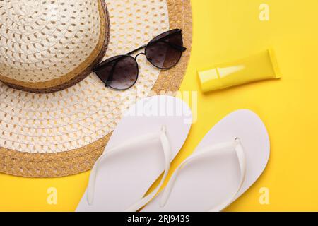 Cappello di paglia, occhiali da sole eleganti, infradito e crema solare su sfondo giallo, piatto. Accessori per la spiaggia Foto Stock