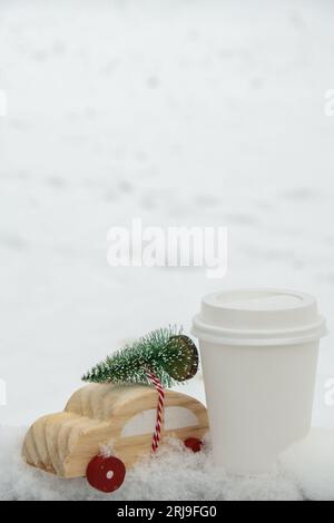 Auto di legno che trasporta l'albero di Natale con la mock della tazza di carta in su del caffè o del cioccolato caldo sopra la neve. Copia spazio per testo Toy auto in paesaggio innevato. Buon Natale e felice anno nuovo concetto. L'inverno sta arrivando biglietto d'auguri Foto Stock