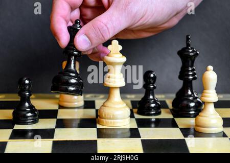 Mano maschile che tiene in mano la regina nera per sconfiggere il re bianco su una scacchiera su sfondo nero. Scacco matto Foto Stock