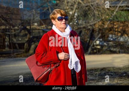 Ritratto di elegante donna di 67 anni in un cappotto rosso brillante con una borsa borgogna, occhiali da sole e sciarpa bianca in una giornata di primavera Foto Stock