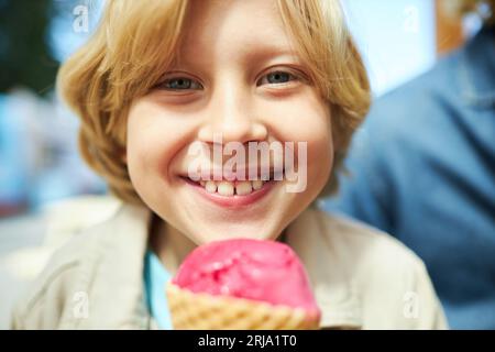 Primo piano di Happy boy che mangia un cono gelato all'aperto al carnevale e sorride alla macchina fotografica Foto Stock