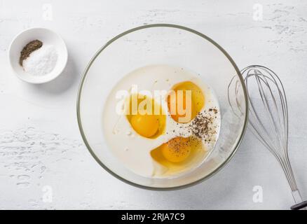 Recipiente di vetro con yogurt, uova e spezie su sfondo azzurro e frusta, vista dall'alto. Impastare l'impasto per la torta, fase di cottura. Foto Stock