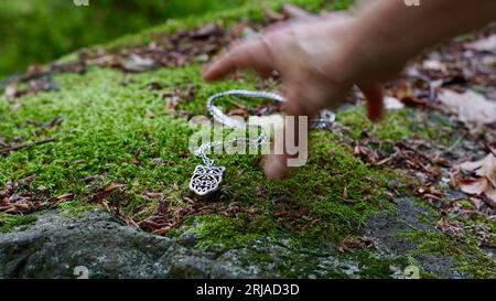 Una mano femminile che cerca una collana d'argento adagiata su una pietra muschiata. Foto Stock