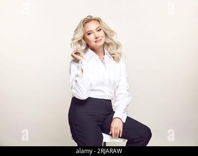 Elegante donna bionda matura che indossa abiti casual e alla moda siede sulla sedia isolata Foto Stock
