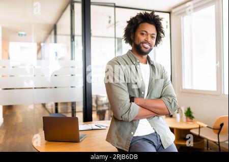 Imprenditore afroamericano riccio di successo si trova in piedi e guarda la macchina fotografica con le braccia piegate, un libero professionista positivo e sorridente in camicia casual nello spazio di coworking, ceo, manager, proprietario di una Start-up Foto Stock