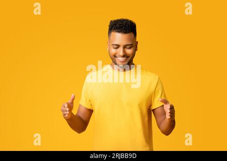 Ritratto di un giovane indiano sorridente in maglietta isolata su sfondo giallo che tiene e guarda lo spazio vuoto, mostra la misura delle dimensioni con le mani, copia l'immagine mockup dello spazio Foto Stock