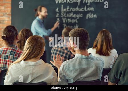 Uomo d'affari serio, il leader risponde alle domande degli ascoltatori che soggiornano alle scrivanie in ufficio con interni in loft. Pubblico nella sala conferenze. Foto Stock