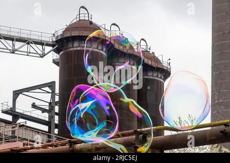 Vista angolare bassa di alcune grandi bolle di sapone colorate di forma sferica che galleggiano di fronte ad alcune torri abbandonate e fatiscenti di acciaio industriale Foto Stock