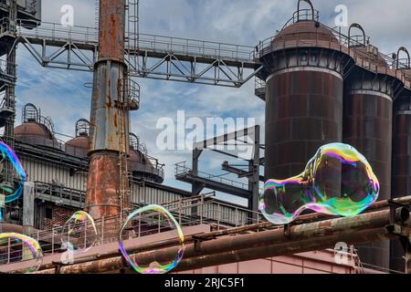 Vista angolare bassa di alcune grandi bolle di sapone colorate di forma sferica che galleggiano di fronte ad alcune torri abbandonate e fatiscenti di acciaio industriale Foto Stock