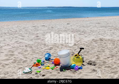 Vista di molti giocattoli da spiaggia nella sabbia, come paletta di sabbia, secchio, rete, muffa da spiaggia altrimenti vuota con mare blu e cielo blu sullo sfondo Foto Stock