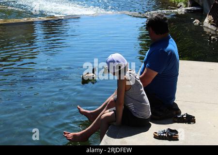 Father and son relax by the banks of the River Sorgue on a warm spring day in  L'Isle sur la Sorgue. Concept for locals relaxing, tourists cooling off Stock Photo