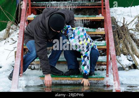 Uomo e bambino che si piegano dalla scala provando acqua ghiacciata con le dita durante la festa Epifania, fiume. Kiev, Ucraina. Foto Stock