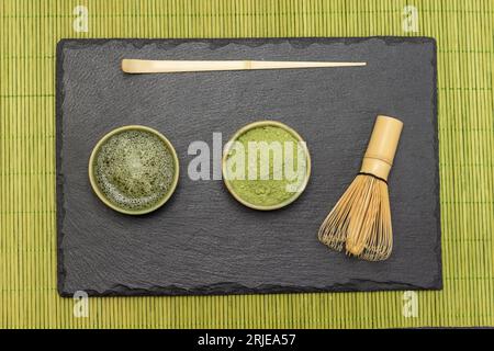 Cucchiaio da tè, frusta di bambù, tè matcha in polvere e tè matcha su tavola. Spianatura piatta. Sfondo di piccoli bastoncini di legno verdi Foto Stock