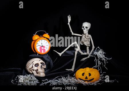 Halloween. Un giocattolo Skeleton che striscia fuori dalla tomba, e agitando la mano, saluta, zucche con le grimaces, un orologio arancione, un cranio su un bac nero Foto Stock