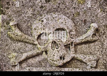 Cranio scolpito e ossa crociate, un antico simbolo di mortalità, su una lapide medievale ricoperta di licheni e intemperie. Foto Stock