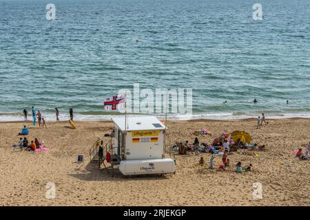 Boscombe, Regno Unito - 11 agosto 2023: Persone sulla spiaggia intorno alla bandiera che volano sopra un posto di guardia Llifeguard RNLI. Foto Stock