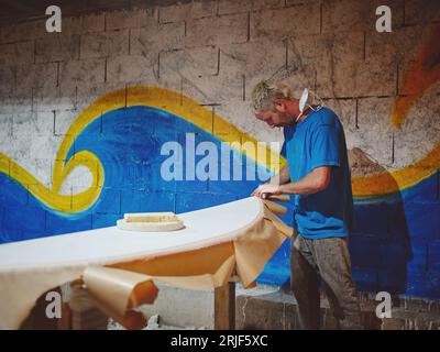 Vista laterale della carta per incollaggio di carpentiere uomo concentrata sui bordi della tavola da surf preparazione per la colorazione in officina Foto Stock