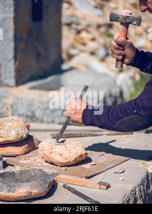 Ritaglia anonimi artigiani che tagliano la roccia con scalpello e martello mentre crei oggetti artigianali in pietra sul banco di lavoro nelle giornate di sole Foto Stock