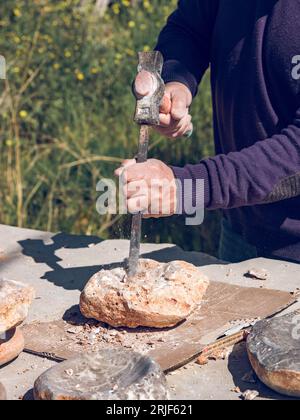 Dalla parte superiore del raccolto, anticamente irriconoscibile, il taglio artigianale di rocce solide con martello e scalpello mentre si fa l'artigianato della pietra in officina all'esterno Foto Stock