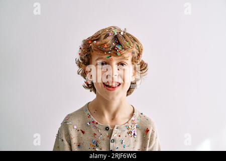 Allegro e grazioso ragazzo biondo dai capelli ricci ricoperto di coriandoli colorati e scintillanti su sfondo bianco Foto Stock