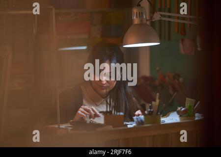 Concentrata orafa etnica adulta con lunghi capelli scuri che esaminano i gioielli mentre si siede al banco da lavoro con vari strumenti e lampada luminosa a stu Foto Stock