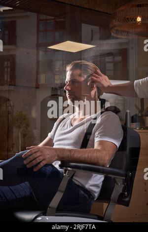 Hipster maschio sicuro di sé seduto in poltrona e ottenere un nuovo taglio di capelli da un parrucchiere professionista nel barbiere Foto Stock