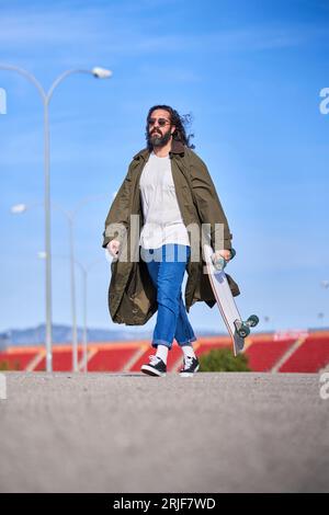 Adulto a pieno corpo, uomo barbato alla moda con occhiali da sole che indossa un elegante impermeabile e jeans che passeggia su strada asfaltata con longboard nelle giornate di sole Foto Stock